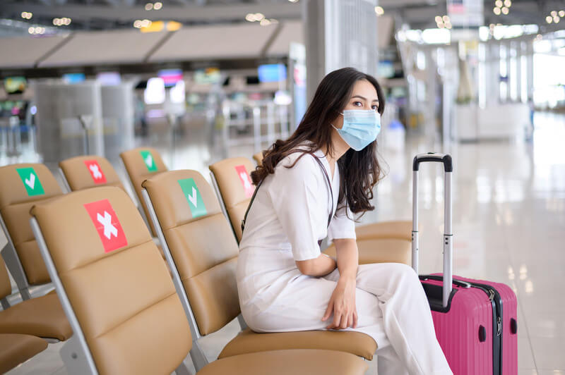 vrouw met mondkapje zit op stoel op vliegveld met een koffer naast haar