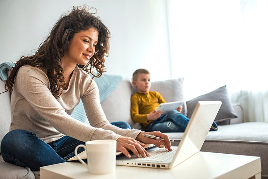 Vrouw werkt thuis achter laptop terwijl haar kind naast haar op de bank zit